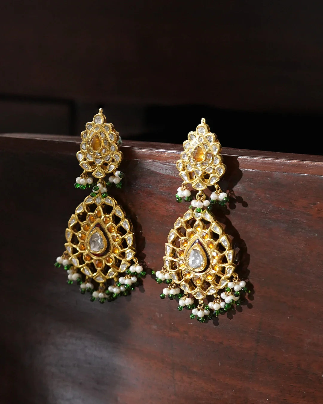 Indus handcrafted gold earrings featuring Polki diamonds, tourmalines, traditional kundan jadai, red meena chudiwork, seed pearls, and green enamel bead fringes.